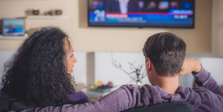 A woman with dark curly hair sitting next to a man on a sofa, they're watching TV. We see the backs of their heads as they speak to each other. They're watching a news program.