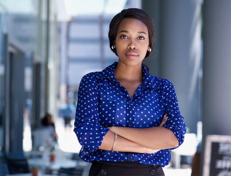 A professional woman with a blue spotted top stands in front of a business/cafe setting, she's crossing her arms across her torso, wearing gold earrings and looking directly at camera.