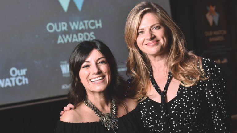 Two public figures Angela Pippos and Julie Zemiro stand close together in front of a projector screen with the Our Watch Awards logo displayed. They are both looking at the camera and smiling.
