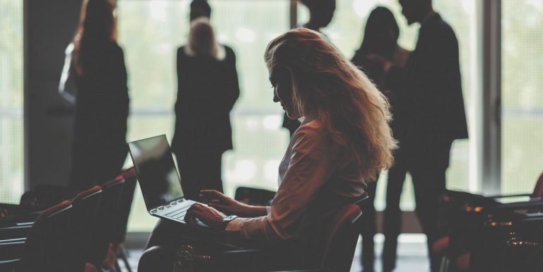 Shillouetted woman sitting in conference room looking at her laptop. She has long hair and there a people standing around talking in the background.