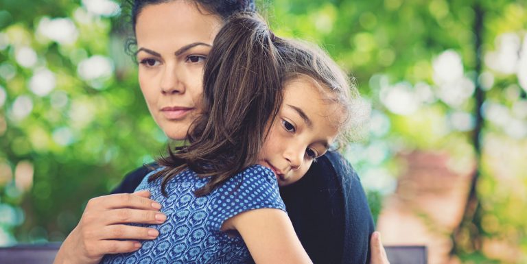 A dark haired woman and a girl embrace each other. There is a greenery in the background.