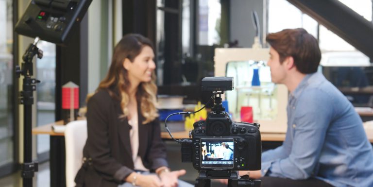 Shallow depth of field image of a young man and woman sitting and talking, interview style, with a camera focussed on them. The woman is speaking.