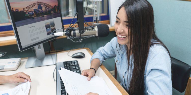 Young woman sits smiling at a radio broadcast desk with a computer screen in the background. She is reading from a piece of paper and sitting opposite a person whose hands we can see on the desk.